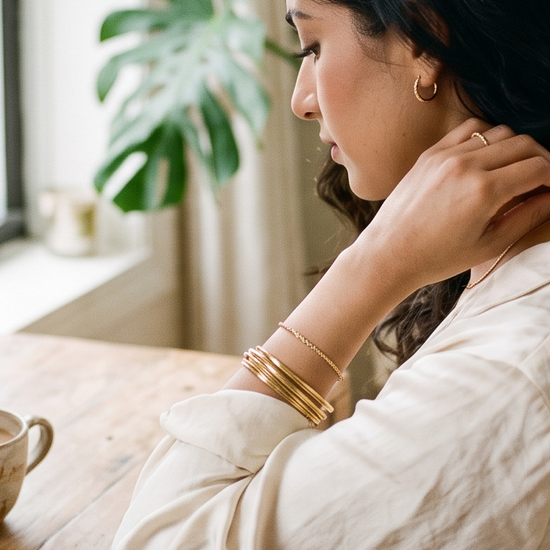 South Asian American woman in a cream blouse fastening a delicate necklace at a sunlit dressing table with gold bangles, chai, and wedding invitations nearby